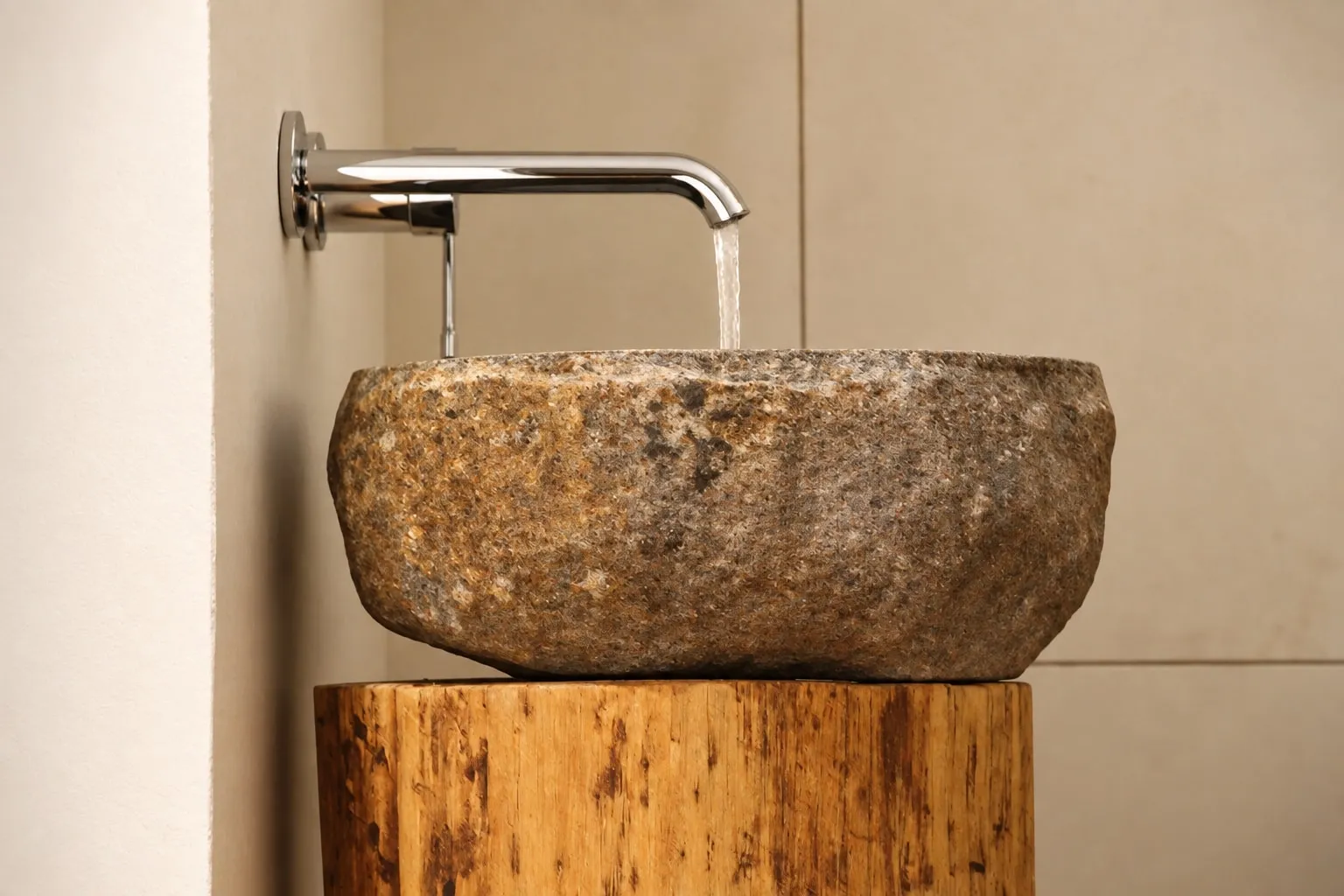 Lavabo ricavato da una pietra grezza appoggiato su un ceppo di legno, con rubinetto cromato a parete da cui scorre l’acqua.