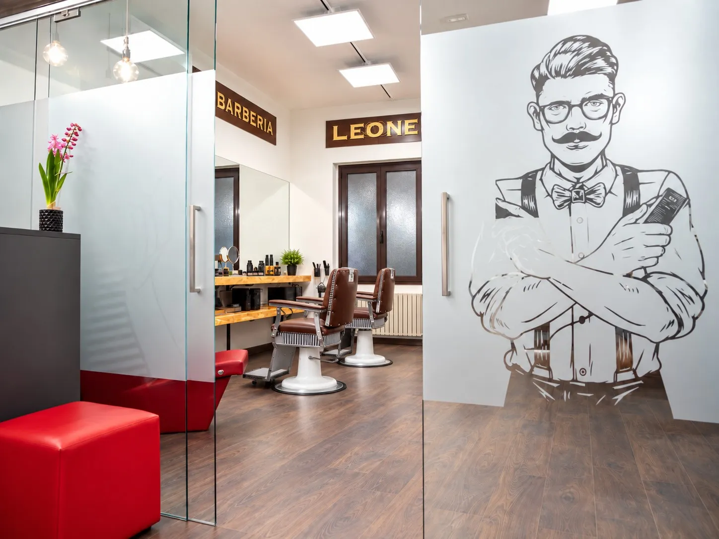 Porta scorrevole in vetro satinato con il disegno di un barbiere con baffi e bretelle; oltre il vetro la sala barbiere con due poltrone in pelle marrone e le insegne Barberia e Leone, in primo piano un pouf rosso