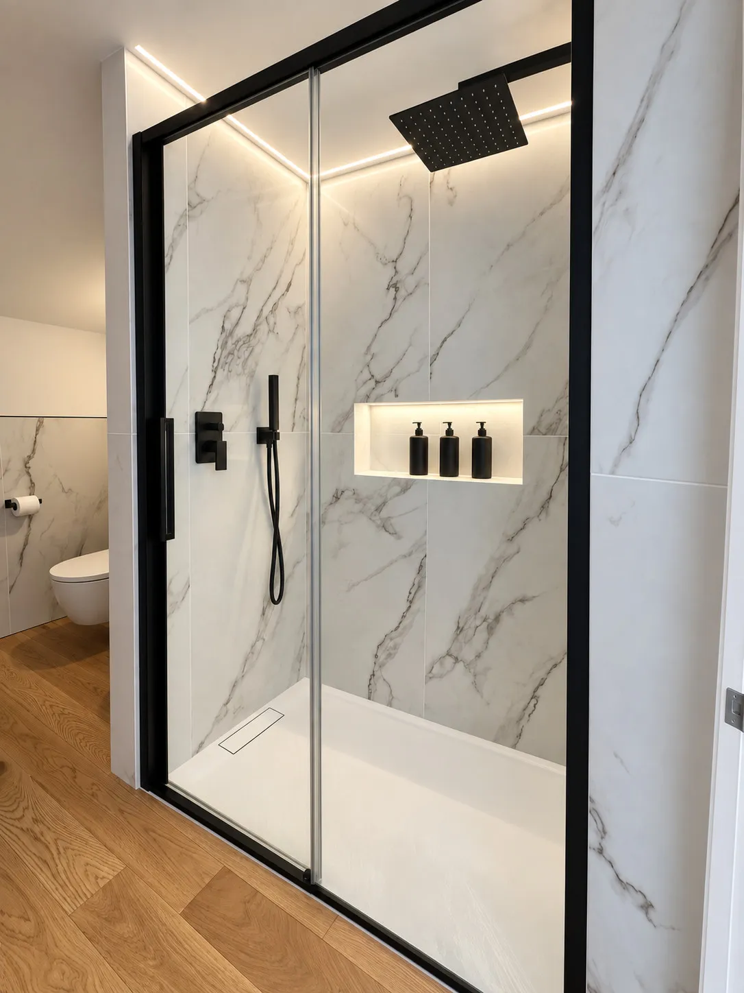 Doccia con profili neri, soffione quadrato e nicchia illuminata fra lastre effetto marmo; sullo sfondo il wc sospeso e il parquet