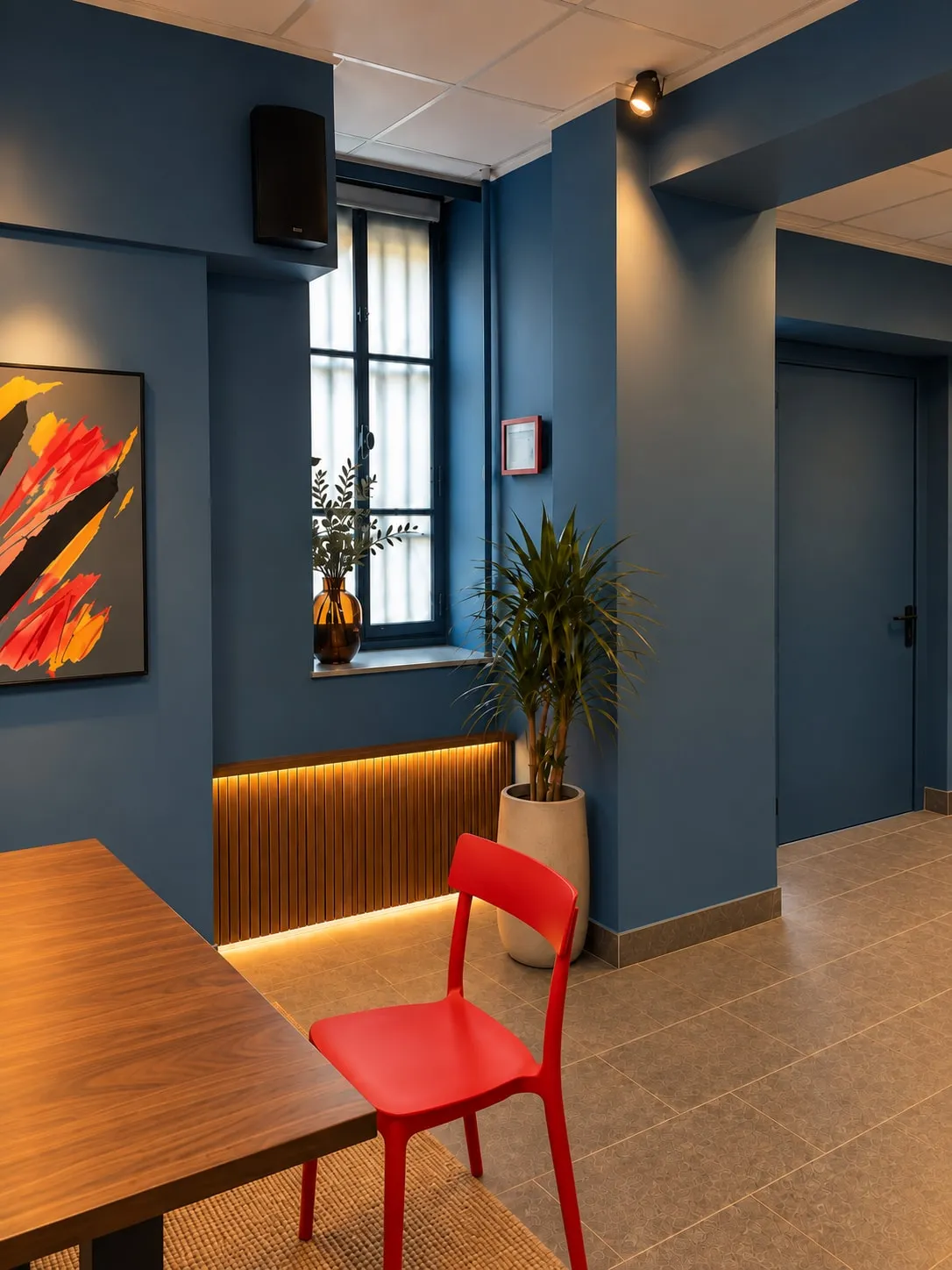 Angolo con finestra: copritermosifone a listelli in legno illuminato dal basso, pianta in vaso, quadro astratto, tavolo in legno e sedia rossa