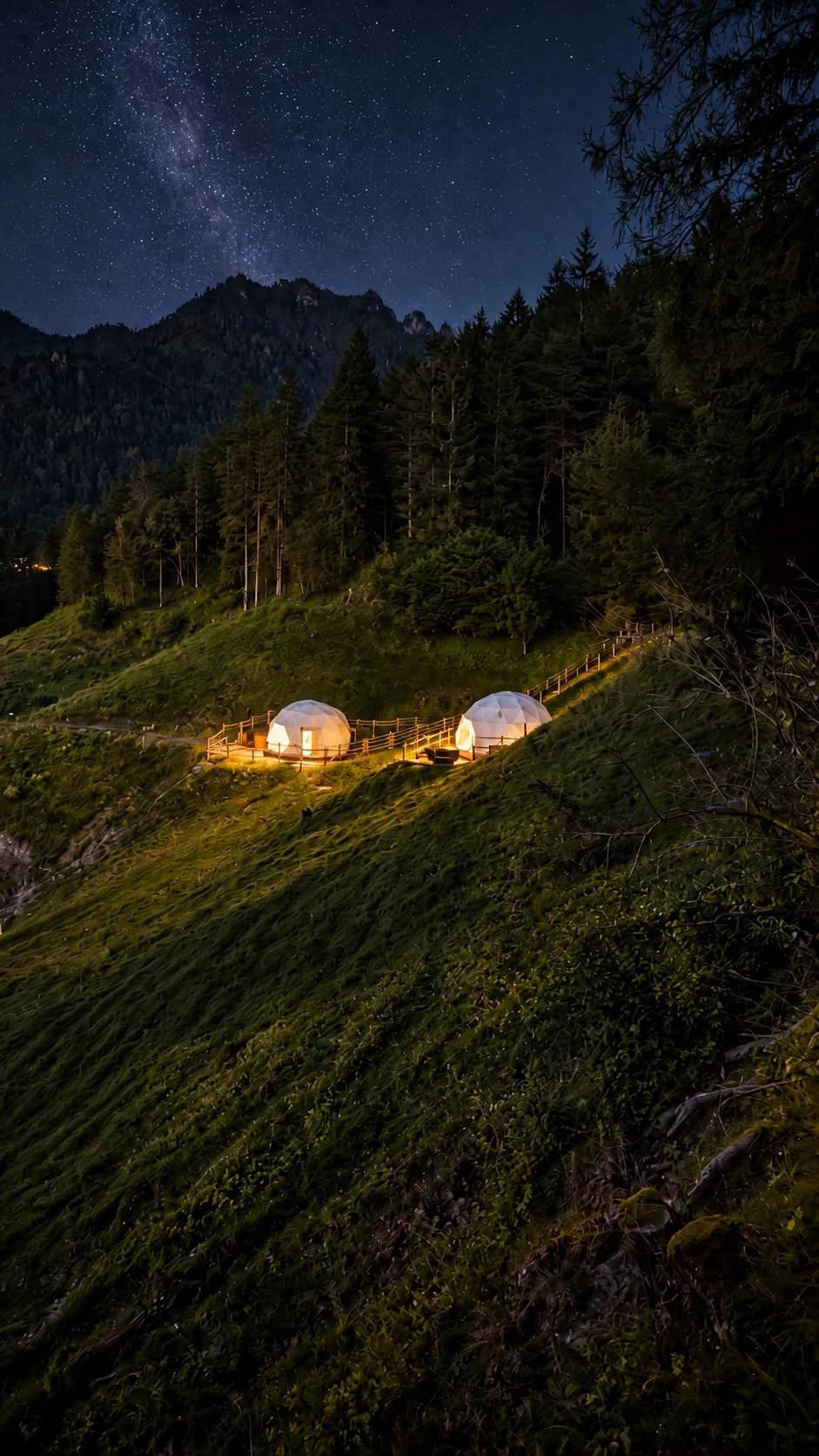 Due lodge geodetici illuminati sul pendio di notte, con le staccionate accese, sotto il cielo stellato e le montagne