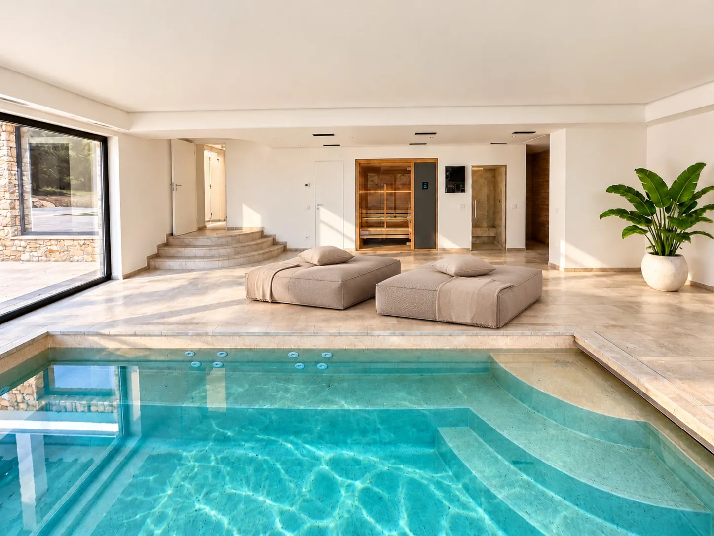 Piscina interna di giorno con acqua azzurra e gradini ad angolo; sul fondo due sedute imbottite, la sauna con porta in vetro e una grande vetrata sul giardino.