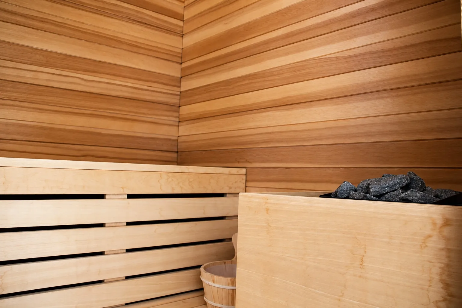 Interno della sauna rivestito in doghe di legno, con la panca chiara a listelli, il secchio di legno e le pietre della stufa.
