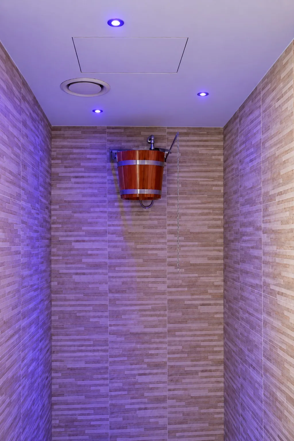 Doccia rivestita in listelli chiari, illuminata di luce viola, con un secchio di legno appeso a parete per la doccia fredda.